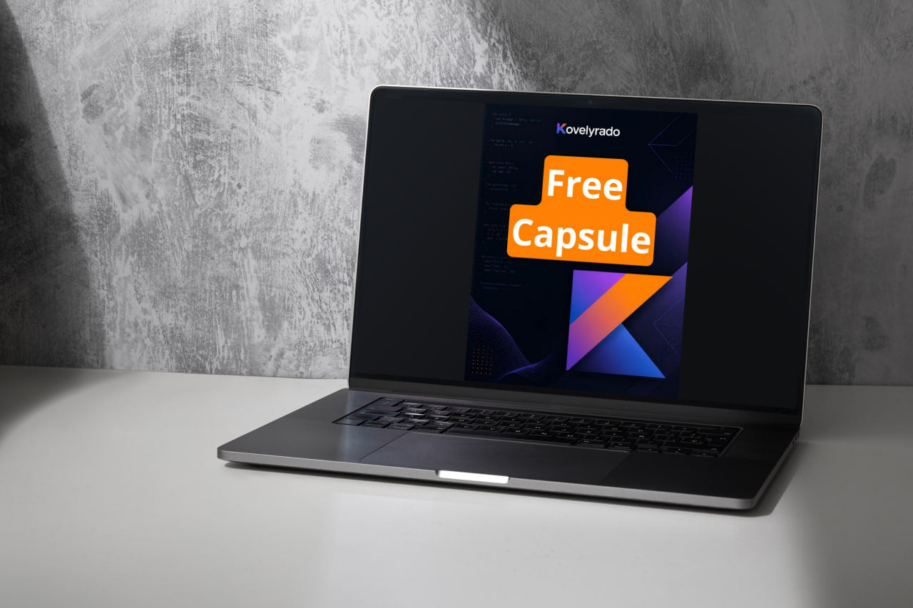 An open laptop sits on a white tabletop against a textured gray wall. The screen displays a promotional graphic with the text 'Kovelrado Free Capsule' in orange and white over a dark background with geometric shapes.