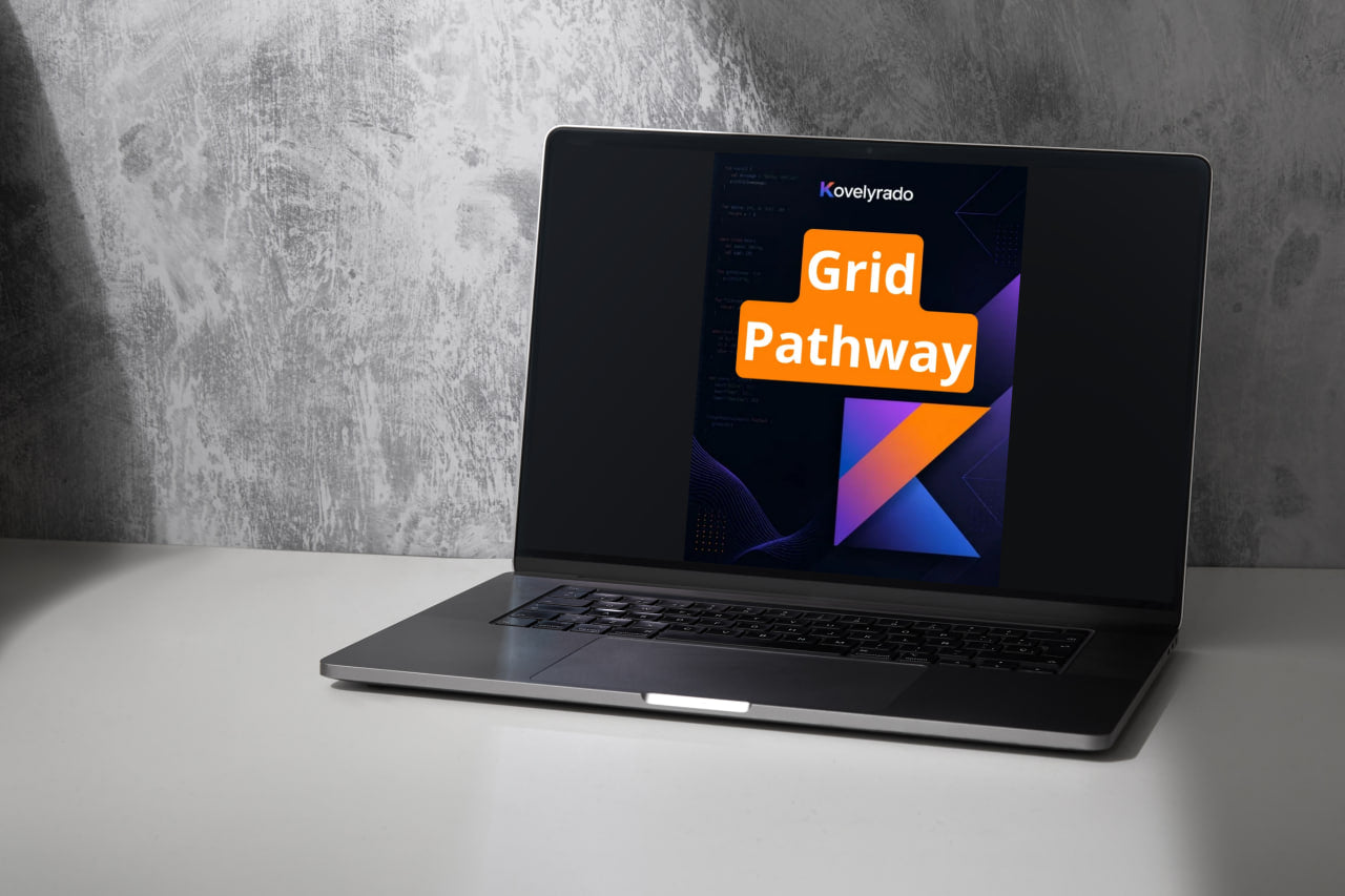 An open laptop sits on a white tabletop against a textured gray wall. The laptop screen displays a digital graphic with the text 'Koveltyado Grid Pathway' in orange and white over a dark background with geometric shapes.