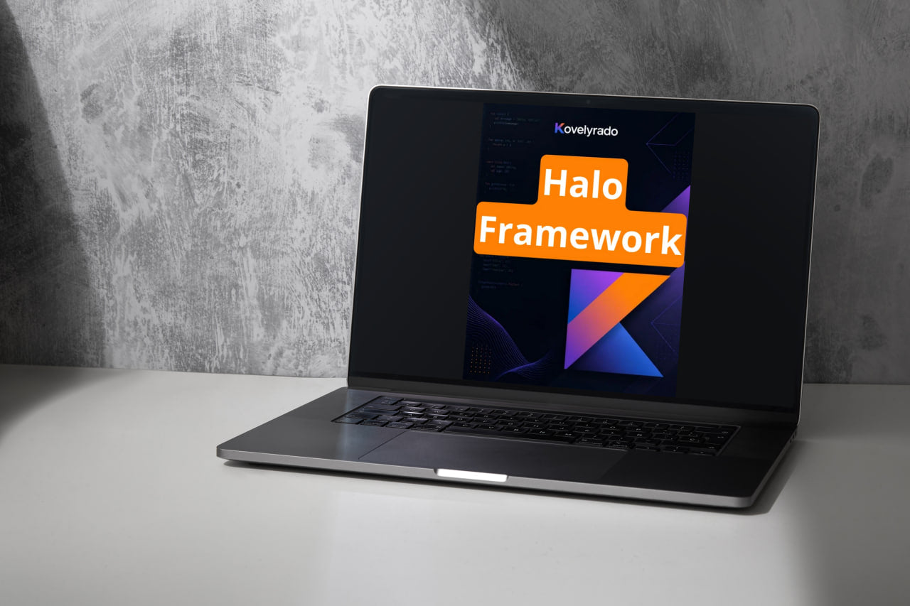 A silver laptop sits open on a white tabletop against a textured gray wall, displaying a screen with the text 'Kovelrado Halo Framework' in orange and white.