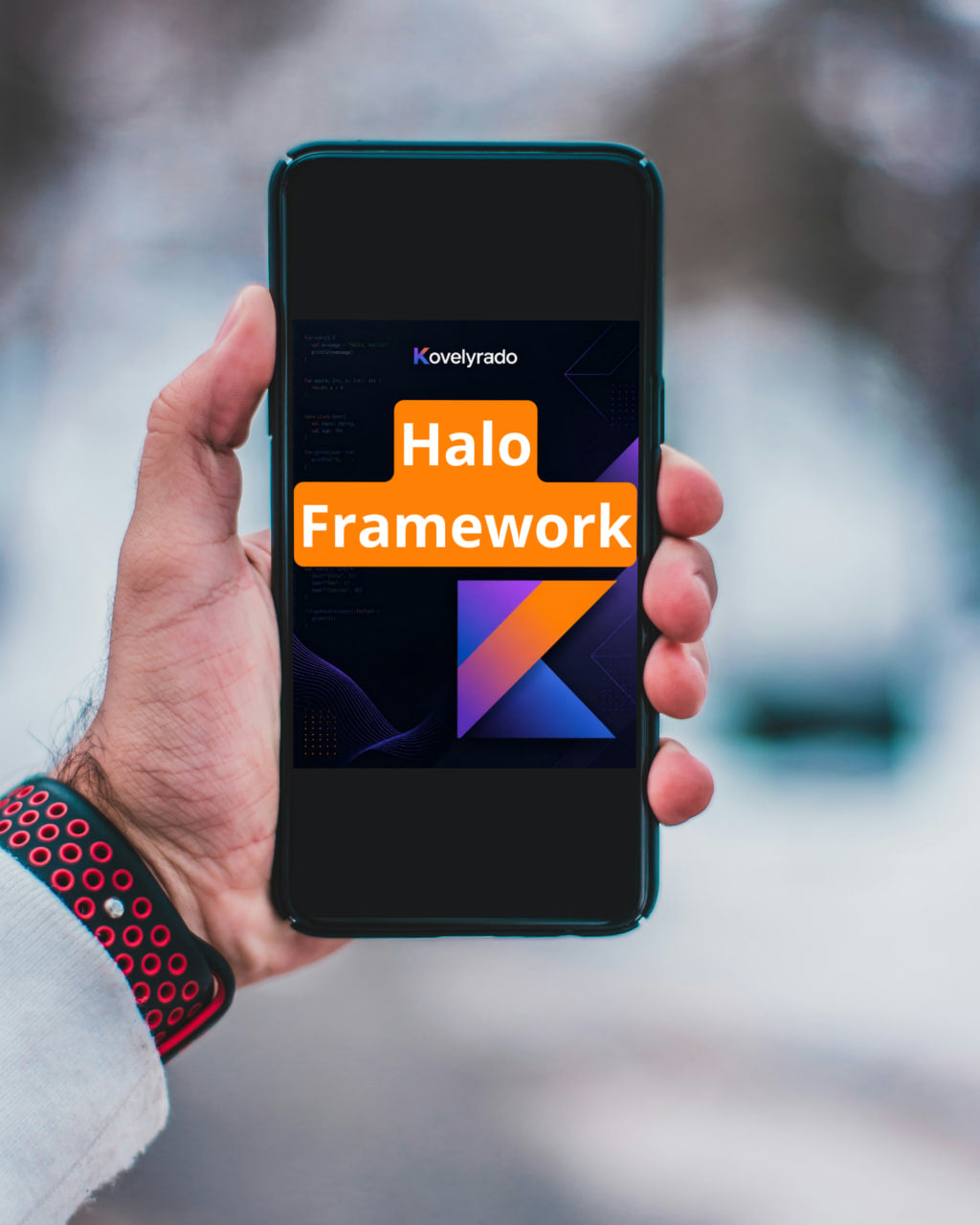 A hand holding a smartphone with a dark screen displaying the text 'Kovelyrado Halo Framework' in orange and white, with a blurred outdoor snowy background.