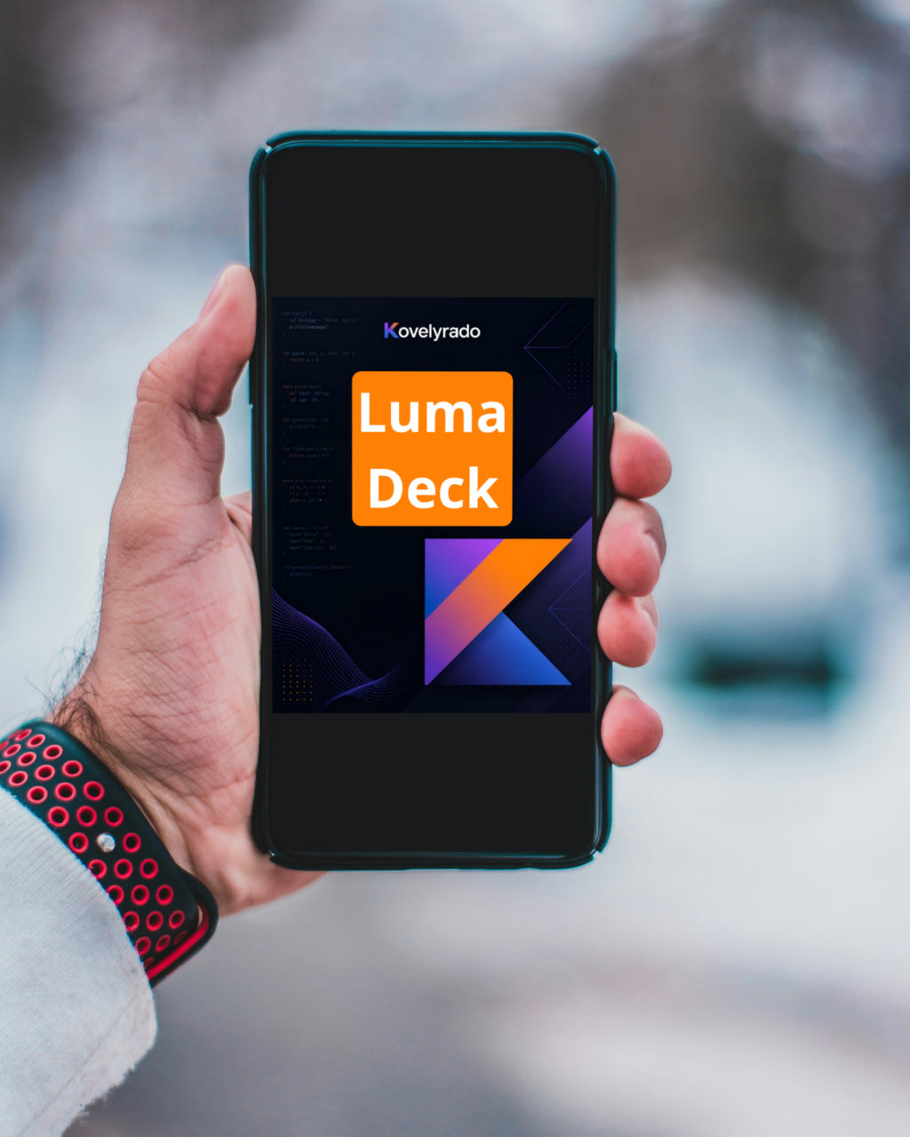 A hand holds a smartphone displaying the Kovyrlado Luma Deck app interface against a blurred outdoor winter background.