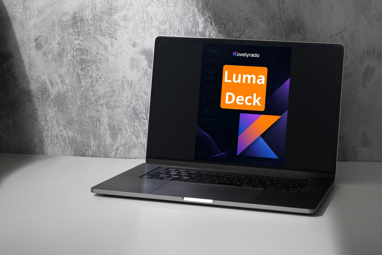 A silver laptop sits open on a white tabletop against a textured gray wall. The screen displays the Kovielyrado Luma Deck interface.